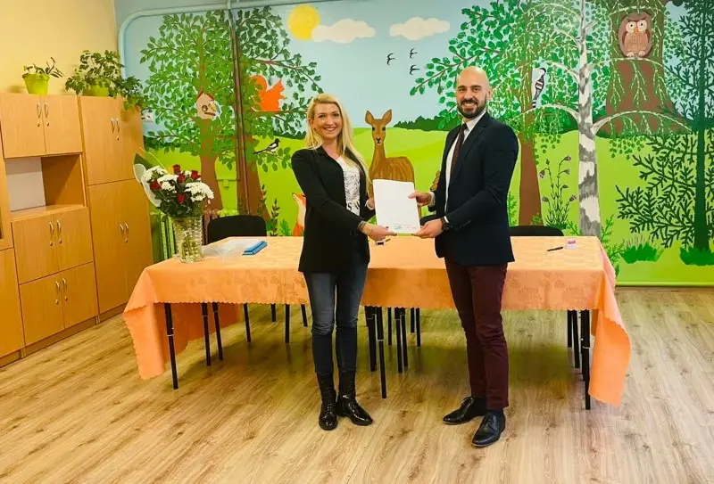 Zdjęcie o orientacji poziomej przedstawia kobietę i mężczyznę stojących wewnątrz sali przedszkolnej, którzy wspólnie trzymają biały dokument. Kobieta po lewej stronie ma na sobie czarną marynarkę, dżinsy i czarne botki, natomiast mężczyzna po prawej jest ubrany w ciemną marynarkę, koszulę z krawatem oraz bordowe spodnie. Przed nimi znajduje się stół przykryty brzoskwiniowym obrusem, na którym stoi wazon z kwiatami. W tle widoczny jest kolorowy mural przedstawiający las z dzikimi zwierzętami, takimi jak sarna, sowa i wiewiórka, co nadaje wnętrzu edukacyjny charakter. Po lewej stronie kadru ustawione są wysokie, drewniane szafki, a na podłodze leży jasny parkiet w odcieniu drewna. Całość kompozycji jest symetryczna i dobrze oświetlona, co podkreśla oficjalny, lecz przyjazny charakter spotkania.