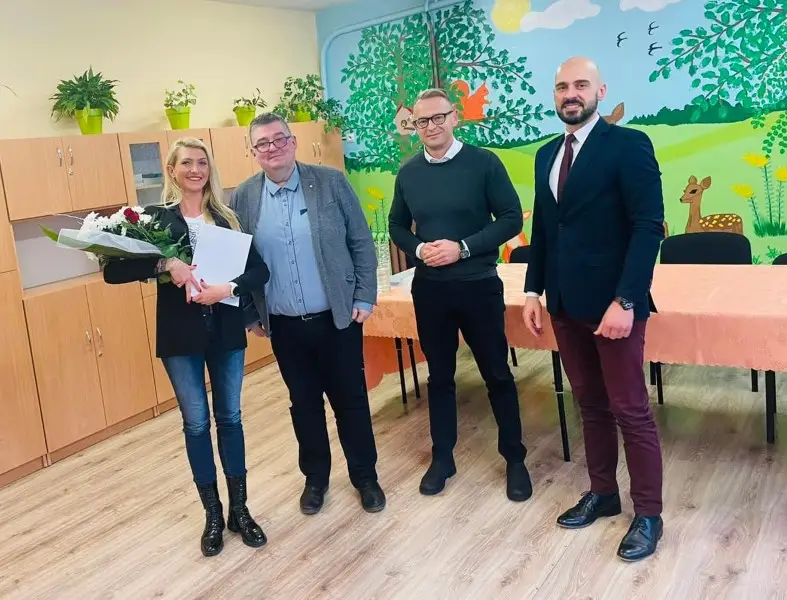 Zdjęcie wykonane w orientacji pionowej przedstawia grupę czterech osób stojących w jasnej sali przedszkolnej lub szkolnej. Po lewej stronie stoi kobieta w czarnej marynarce i dżinsach, trzymająca bukiet kwiatów oraz białą teczkę. Obok niej znajdują się trzej mężczyźni: jeden w szarej marynarce i koszuli, drugi w ciemnozielonym swetrze, a trzeci w eleganckiej granatowej marynarce i bordowych spodniach. Tło pomieszczenia zdobi kolorowy mural przedstawiający las, zwierzęta i słońce, a przy ścianie ustawiono drewniane szafki z roślinami doniczkowymi. Kompozycja obrazu jest symetryczna, a postacie znajdują się w centrum kadru, co sugeruje formalne spotkanie lub uroczyste wręczenie dokumentów. Całość utrzymana jest w jasnej, przyjaznej kolorystyce typowej dla placówek edukacyjnych.