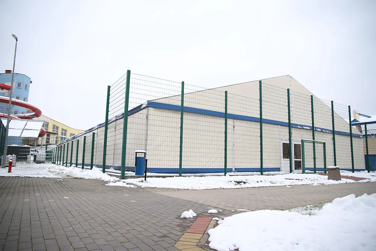 Zdjęcie przedstawia dużą halę namiotową o jasnych ścianach i dwuspadowym dachu, ogrodzoną wysokim zielonym płotem z siatki stalowej. Obiekt o proporcjach zbliżonych do 3:2 znajduje się na wybrukowanym placu pokrytym częściowo płatami śniegu, co sugeruje zimową aurę. Po lewej stronie widać fragment kolorowej zjeżdżalni wodnej oraz nowoczesną latarnię, a na pierwszym planie ustawiony jest niebieski kosz na śmieci. Hala posiada wyraźne niebieskie obramowanie konstrukcyjne oraz podwójne przeszklone drzwi wejściowe, które są centralnym punktem prawej ściany. Całość kompozycji zachowana jest w chłodnej tonacji, gdzie dominującym elementem jest jasna bryła hali kontrastująca z szarością nieba.